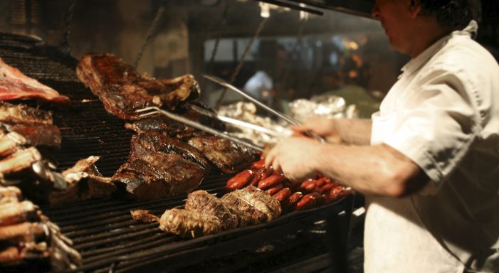 A person using tongs to grill various meats over an open fire.